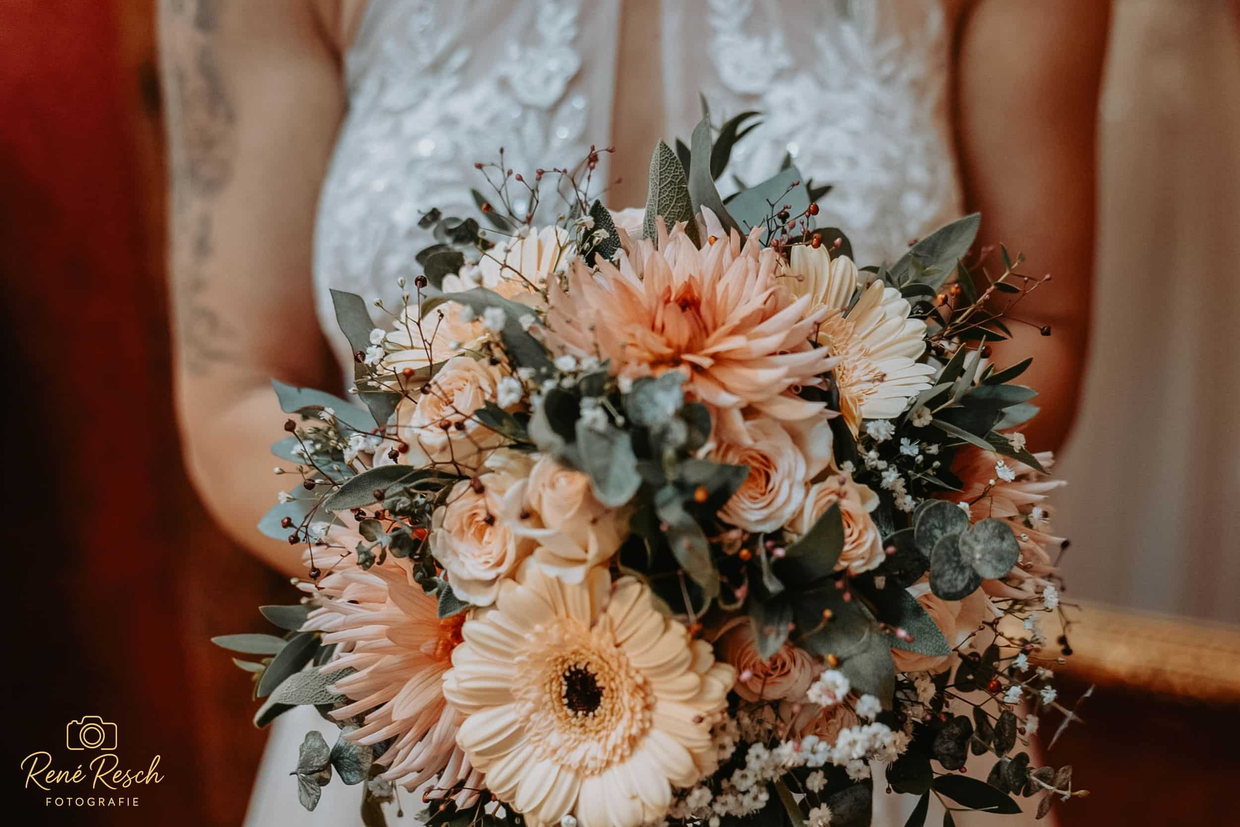 Nahaufnahme einer Braut, die einen Strauß aus blassrosa und weißen Blumen mit Grünzeug vor ihrem Spitzenhochzeitskleid hält. Der Strauß enthält Gerbera-Gänseblümchen und andere zarte Blüten.