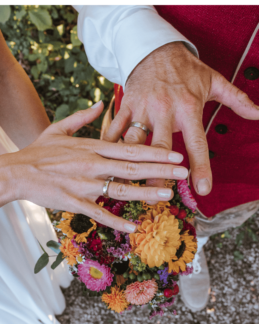 Zwei Personen zeigen ihre Hände mit Eheringen und legen sie über einen bunten Blumenstrauß. Eine Person trägt einen weißen Ärmel, die andere eine rote Weste. Die Szene deutet auf eine Hochzeit oder eine Feier im Freien hin.