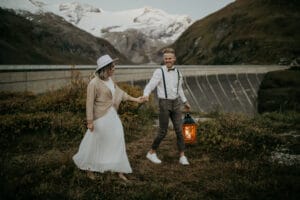 Ein Paar in legerer Hochzeitskleidung geht Hand in Hand durch eine grasbewachsene Landschaft in der Nähe eines großen Staudamms und träumt von seiner perfekten Hochzeit. Die Person rechts hält eine Laterne. Schneebedeckte Berge und Grünflächen bieten eine atemberaubende Kulisse, während sie über ihre Hochzeitsplanung nachdenken.