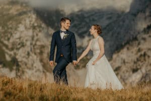 Braut und Bräutigam tauschen liebevolle Blicke aus, ihre Finger sind ineinander verschränkt, auf einem grasbewachsenen Hügel mit majestätischen Bergen im Hintergrund. In ihrem eleganten weißen Kleid und seinem eleganten dunklen Anzug verkörpern sie die Freude einer Hochzeit inmitten der Pracht der Natur.