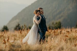 Ein Paar steht auf einer Wiese mit Bergen im Hintergrund. Die Frau im hellen Kleid umarmt den Mann im dunklen Anzug von hinten. Diese ruhige, naturnahe Umgebung fängt die Essenz einer Hochzeit wunderschön ein und weckt Romantik und Hochzeitsträume.
