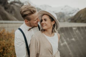 Ein Paar steht eng beieinander und lächelt sich an seinem Hochzeitstag an. Der Mann trägt ein weißes Hemd mit Hosenträgern und Fliege, während die Frau ein weißes Kleid und einen Hut trägt. Sie sind im Freien, mit Bergen im Hintergrund, die wunderschön verschwommen sind.
