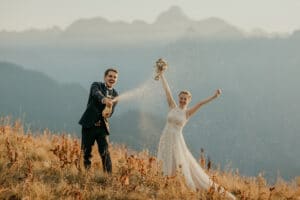 Ein Paar feiert seine Hochzeit auf einem Berghang, während der Mann eine Champagnerflasche öffnet und sie in die Luft sprüht. Die Frau hält einen Blumenstrauß und streckt freudig die Arme in die Höhe. Sie sind umgeben von hohem Gras und entfernten Berggipfeln, die die Essenz ihres perfekten Tages einfangen.