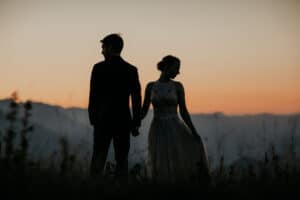 Die Silhouette eines Paares steht bei Sonnenuntergang Hand in Hand auf einem grasbewachsenen Hügel. Der perfekte Moment, um die Hochzeit zu planen. Der Himmel ist in Orange und Blau gehalten, im Hintergrund sind Berge zu sehen. Die Frau trägt ein fließendes Kleid und der Mann einen Anzug, der Eleganz und Romantik verkörpert.