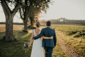 Ein Paar im Hochzeitskleid geht bei Sonnenuntergang Arm in Arm einen von Bäumen gesäumten Weg entlang und verkörpert damit die Essenz einer Hochzeit. Die Braut, die ihren Blumenstrauß in einem weißen Kleid umklammert, und der Bräutigam in seinem Anzug genießen den warmen Schein der Sonne über der ruhigen Landschaft.