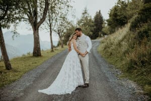 Braut und Bräutigam umarmen sich auf einem ländlichen Schotterweg, der von Bäumen und Grün gesäumt ist und ihrer Hochzeit einen Hauch von rustikalem Charme verleiht. Die Braut trägt ein weißes Spitzenkleid, während der Bräutigam einen hellen Anzug trägt. Sanfte Hügel und ein bewölkter Himmel verstärken diese ruhige und romantische Hochzeitsatmosphäre.