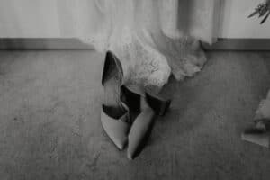 Ein Schwarzweißbild zeigt ein Paar Schuhe mit Absatz auf einem strukturierten Boden neben einem Spitzenstoffvorhang – eine perfekte Verkörperung der Eleganz von Hochzeitsmomenten.