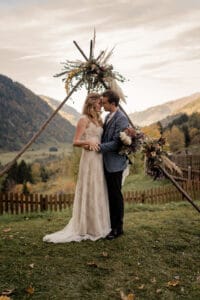 Braut und Bräutigam stehen eng beieinander und umarmen sich vor einem rustikalen Blumenbogen. Die malerische Bergkulisse verleiht ihrer Hochzeit mit einem Holzzaun und Herbstlaub unter einem bewölkten Himmel Magie – eine idyllische Kulisse, die sich perfekt für jede Hochzeitsplanung eignet.