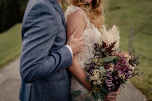 Ein Paar steht eng beieinander, wobei der Mann im blauen Anzug die Frau im Spitzenkleid, die einen Strauß Wildblumen trägt, sanft hält. Sie stehen draußen auf einem Weg mit viel Grün im Hintergrund und halten einen besonderen Moment ihrer Hochzeitsplanung fest.