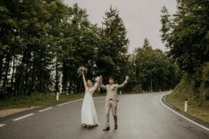 Ein glückliches Paar, das in seinem Hochzeitsglück schwelgt, geht Hand in Hand eine nasse, kurvenreiche Straße entlang, die von üppigen grünen Bäumen umgeben ist. Die Braut hält einen Blumenstrauß hoch und beide lächeln und haben die Arme feierlich in die Höhe gestreckt.