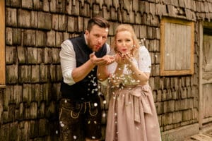 Ein Mann und eine Frau in traditioneller bayerischer Kleidung, die wahrscheinlich ihre Hochzeit planen, pusten Konfetti in Richtung Kamera, während sie vor einem rustikalen Holzgebäude stehen. Der Mann trägt Lederhosen und die Frau ein Dirndl. Beide wirken fröhlich.