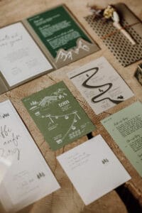 Auf einem Holztisch wird elegant eine Auswahl an Hochzeitspapeterie präsentiert – ein wesentlicher Bestandteil der Hochzeitsplanung. Grüne und weiße Einladungen, Karten und handschriftliche Notizen sind mit Abbildungen von Bergen und Bäumen geschmückt. Ein Arrangement aus getrockneten Blumen ist teilweise sichtbar.