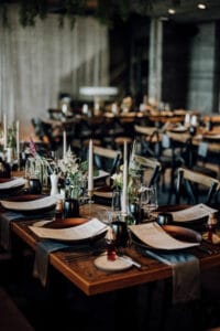 Ein wunderschön gedeckter Esstisch mit elegantem Gedeck, perfekt für eine Hochzeit, mit Kerzen und Blumenschmuck in einem schwach beleuchteten Restaurant. Holzstühle und zusätzliche Tische schaffen eine gemütliche Atmosphäre, ideal für diejenigen, die gerade mitten in der Hochzeitsplanung stecken.