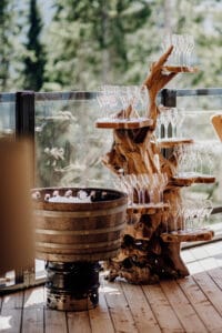 Eine Ausstellung leerer Weingläser, perfekt für eine Hochzeitsfeier, ist auf einem baumartigen Holzständer neben einem mit Eis und Flaschen gefüllten Fass arrangiert, auf einem Holzdeck mit Glasgeländer und üppigen Bäumen im Hintergrund.