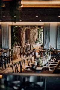 Ein gemütliches Restaurant-Interieur, perfekt für eine Hochzeit, mit Holzdekor und eleganten Tischen zum Essen. Gepolsterte Stühle und gedämpftes Licht schaffen eine warme Atmosphäre, während grüne Akzente und ein Spiegel die natürliche Ästhetik widerspiegeln – ideal für alle, die heiraten möchten.