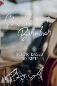 Das Bild zeigt eine Glastür mit elegantem Schriftzug „Matthias & Bernhard“ und fettgedruckten Buchstaben mit der Aufschrift „SCHEN, DASS’D DO BIST!“. Rechts ist eine Andeutung einer Person zu sehen und darunter befindet sich eine Berggrafik. Diese Szene könnte Teil einer exquisiten Hochzeitslocation sein.