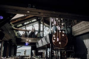 Ein modernes, schwach beleuchtetes Restaurant-Interieur mit großen Fenstern und Holzbalken, perfekt für Hochzeitsplanung. Eine glänzende Discokugel hängt in der Nähe des Barbereichs und sorgt für festliche Stimmung. Der Sitzbereich im zweiten Stock ist ideal für eine Hochzeitsfeier, bei der die Gäste unter einem Kronleuchter mit baumelnden Lichtern plaudern.