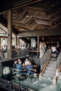 Ein modernes Restaurant mit Holzdecken und großen Fenstern sowie mehreren Essebenen ist ideal für alle, die eine Hochzeitsfeier planen. Die Gäste sitzen an verschiedenen Tischen und genießen Mahlzeiten und Gespräche. Sanfte Beleuchtung und eine gemütliche Atmosphäre runden das Ambiente ab.