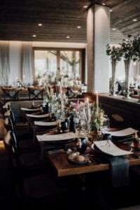 Ein warm beleuchteter Speisesaal, ideal für eine Hochzeitsfeier, bietet elegant gedeckte Tische mit Kerzen, Blumen und Menüs auf Holzoberflächen. Der Raum strahlt eine gemütliche Atmosphäre mit sanfter Beleuchtung und einer Mischung aus modernen und rustikalen Dekorelementen aus, perfekt für Ihren besonderen Tag.