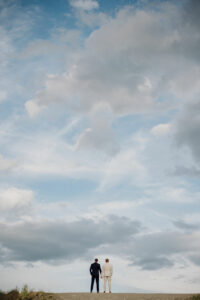 Zwei Menschen stehen nebeneinander auf einem Hügel und blicken in einen weiten Himmel voller dramatischer Wolken. Während sie über ihre gemeinsame Zukunft nachdenken, deuten ihre passenden Anzüge auf eine bevorstehende Hochzeit hin und symbolisieren Offenheit und Planung unter dem weiten Himmel.