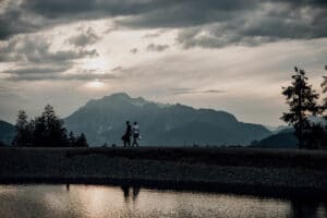 Ein Paar geht bei Sonnenuntergang einen Bergrücken entlang, die Silhouette hebt sich vor einem dramatischen Himmel ab und lässt die Romantik einer Hochzeit aufkommen. Die Szene zeigt majestätische Berge und einen ruhigen See, der gedämpftes Licht reflektiert, während vereinzelte Wolken die ruhige Atmosphäre verstärken – perfekt für Heiraten-Träume.