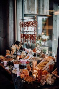 Ein rustikaler Buffettisch, perfekt für Ihre Hochzeit, geschmückt mit Grissini, Crackern, Dips und Käse. Im Hintergrund hängen getrocknete Würste, während brennende Kerzen ein warmes Licht verbreiten. Ein Fenster reflektiert Licht in den gemütlichen Raum und schafft eine bezaubernde Kulisse für Ihren besonderen Tag.