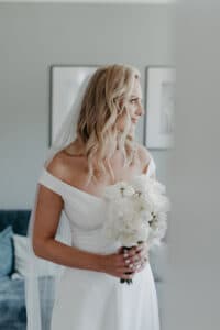 Eine Braut steht in einem sanft beleuchteten Raum und hält einen Strauß weißer Blumen in der Hand. Sie trägt ein schulterfreies weißes Kleid und einen Schleier, ihr langes, welliges blondes Haar fällt ihr über den Rücken. Die Kulisse ist elegant arrangiert – ideal für alle, die sich gerade intensiv mit der Hochzeitsplanung beschäftigen – und Kunstwerke schmücken die hellen Wände.