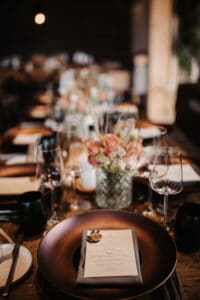 Ein langer Holztisch ist elegant für ein Hochzeitsessen gedeckt, mit Weingläsern, schwarzen Tellern und schwarzen Servietten. Ein Blumengesteck mit rosa und weißen Blumen verläuft in der Mitte des Tisches und schafft eine warme und einladende Atmosphäre, perfekt für Ihre Hochzeitsplanung.