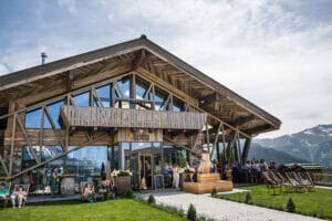 Eine moderne Berghütte aus Holz mit großen Glasfenstern und einem Balkon vor der Kulisse schneebedeckter Gipfel, perfekt für eine malerische Hochzeit. Die Leute versammeln sich draußen, genießen den sonnigen Tag auf einer Wiese mit Holzbänken und Stühlen und halten Erinnerungen an ihren besonderen Hochzeitsmoment fest.