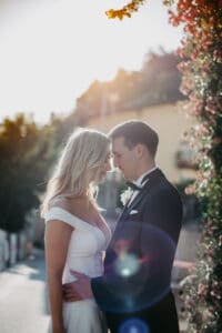 Braut und Bräutigam stehen eng beieinander und umarmen sich in einer sonnenbeschienenen Umgebung im Freien. Die Braut trägt ein weißes Kleid und der Bräutigam einen schwarzen Anzug. Sie stehen in der Nähe von Laub, mit einem Linsenreflexionseffekt der Sonne – und fangen die Essenz des Heiratens inmitten der Schönheit der Natur perfekt ein.
