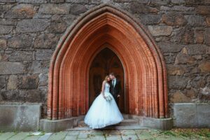 Braut und Bräutigam küssen sich vor dem Eingang einer rustikalen Kirche – perfekt für alle, die von einer malerischen Hochzeit träumen. Die Braut trägt ein weißes Kleid, der Bräutigam einen Anzug. Aufwändige Steinarbeiten zieren den gewölbten Eingang, während gefallene Blätter die romantische Atmosphäre noch verstärken.
