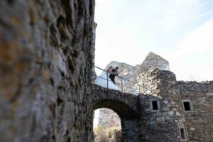 Ein Paar in Hochzeitskleidung steht umarmend auf einer Steinbrücke in einer alten Burgruine, im Hintergrund ein heller Himmel. Die alten Steinmauern verleihen der romantischen Szene einen rustikalen Charme und verkörpern ihren Traum, endlich inmitten solch bezaubernder Geschichte zu heiraten.