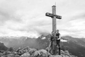 Ein Schwarzweißfoto eines Paares, das neben einem großen Holzkreuz auf einem felsigen Berggipfel steht und die Essenz ihrer Hochzeit einfängt. Die Frau hält einen Blumenstrauß, der ihre Hochzeitsreise inmitten nebliger Berge und Wolken im Hintergrund symbolisiert.