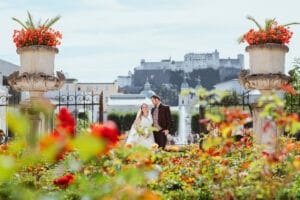 Ein Paar posiert für ein Hochzeitsfoto in einem farbenfrohen Garten voller roter und orangefarbener Blumen. Im Hintergrund sind historische Gebäude und ein Schloss zu sehen, die eine malerische und romantische Szene schaffen.