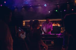 Ein DJ steht an einem Pult unter bunten Bühnenlichtern und mischt Musik für eine Hochzeitsfeier. Im Vordergrund tanzende Menschensilhouetten. Die Atmosphäre ist lebhaft, Rauch und eine lebhafte Menge tragen zur Partystimmung bei.