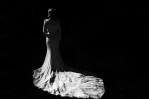 Ein Schwarzweißbild einer Person in einem langen, fließenden Kleid, die im Halbschatten steht, erinnert an die Eleganz einer Hochzeit. Die Schleppe des Kleides ist elegant ausgebreitet und fängt das Licht vor einem dunklen Hintergrund ein, als würde sie einen zeitlosen Moment der Hochzeit festhalten.