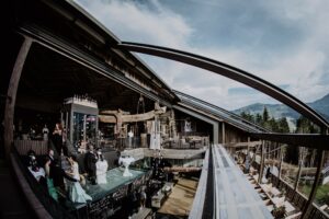 Ein eleganter Innen- und Außenraum mit Holzarchitektur, in dem Gäste bei einem Hochzeitsempfang zu sehen sind. Ein transparenter Glasgang erstreckt sich von einem offenen Bereich und bietet einen malerischen Blick auf die Berge im Hintergrund unter einem teilweise bewölkten Himmel – perfekt für eine sorgfältige Hochzeitsplanung.