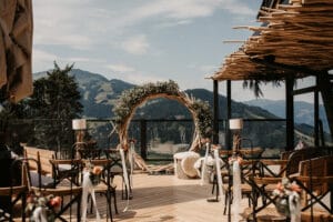 Eine Hochzeit im Freien auf einem Holzdeck bietet einen runden Blumenbogen mit Grünpflanzen, rustikale Holzstühle, die mit Blumen geschmückt sind, und einen atemberaubenden Blick auf die Berge unter einem klaren Himmel. Perfekt für alle, die ihre Traumhochzeit mitten in der Natur planen.