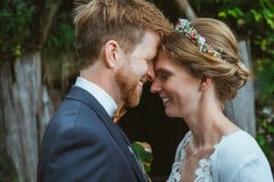 Braut und Bräutigam teilen mitten in ihrer Hochzeit einen intimen, freudigen Moment, berühren sich an der Stirn und lächeln. Die Braut trägt einen Blumenkopfschmuck und ein Spitzenkleid; der Bräutigam trägt einen Anzug mit Fliege. Sie stehen draußen inmitten von leuchtendem Grün.