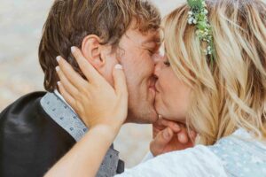 Inmitten der Glückseligkeit ihrer Hochzeit küsst sich ein Paar in atemberaubender Hochzeitskleidung freudig. Die Frau, geschmückt mit einem Blumenhaarschmuck, legt sanft ihre Hände auf das Gesicht des Mannes, beide umarmen sich mit geschlossenen Augen voller Freude und Feierlichkeit.