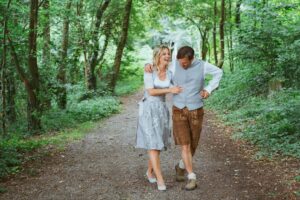 Ein Paar geht glücklich einen Waldweg entlang und schwelgt in seiner Hochzeitsfreude. Die Frau trägt ein hellblaues Kleid und High Heels, während der Mann Lederhosen und eine graue Weste trägt. Sie lachen, umgeben von üppigen grünen Bäumen und Laub.