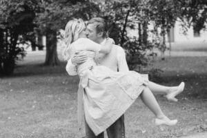 Ein Schwarzweißfoto fängt einen freudigen Moment ein, als ein Mann eine Frau glücklich hochhebt, um ihre Hochzeit zu feiern, und ihr einen Kuss gibt. Sie befinden sich draußen in einer parkähnlichen Umgebung mit Bäumen und Gras im Hintergrund, die Frau trägt elegante High Heels und der Mann Abendgarderobe.