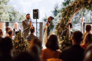 Ein Mann und eine Frau singen bei einer Hochzeitszeremonie in Innenräumen in Mikrofone. Sie stehen neben einem dekorativen Bogen aus Grünpflanzen, während die Gäste sitzen und durch große Fenster den atemberaubenden Blick auf Bäume und Berge genießen.