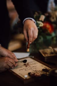 Eine Person versiegelt ein Pergament mit Wachs und verwendet dazu einen Stempel, während eine andere Hand das Papier festhält – ein symbolischer Akt inmitten der heiklen Kunst der Hochzeitsplanung. Der Tisch ist mit Blumenarrangements und Schreibutensilien geschmückt, was auf eine formelle oder zeremonielle Unterzeichnung schließen lässt.