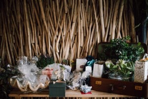 Ein Tisch ist mit verschiedenen Geschenktüten und verpackten Paketen geschmückt und fängt den Geist einer Hochzeitsfeier perfekt ein. Vor einer kunstvollen Holzwand ist die Szene wunderschön mit Grünpflanzen und Blumenarrangements dekoriert, was die festliche Atmosphäre verstärkt.