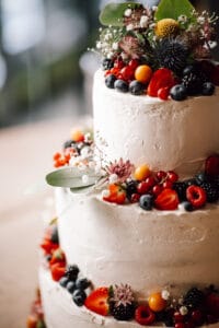 Eine dreistöckige weiße Torte, perfekt für eine Hochzeit, ist mit frischen Beeren wie Erdbeeren, Blaubeeren, Himbeeren und Physalis geschmückt. Mit grünen Blättern und kleinen Blumen dekoriert, ergibt sie eine lebendige und elegante Präsentation, die zu jeder Hochzeitsplanung passt.
