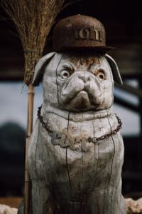 Eine Holzskulptur eines Mopses, perfekt als Hochzeitsplanungsdeko, trägt einen verrosteten Helm mit der Aufschrift „Ton“ und eine Halskette mit dem Namen „Edel“. Auffallend sind seine übertriebenen Gesichtsausdrücke. Auf einer strukturierten Oberfläche lehnt ein Besen dagegen.
