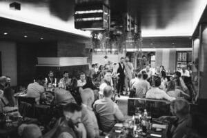 Ein Schwarzweißfoto eines überfüllten Restaurants fängt die lebhafte Atmosphäre ein, die oft für Hochzeitsfeiern gewünscht wird. Gäste sitzen an Tischen, speisen und unterhalten sich, während zwei Personen in der Mitte stehen und inmitten der rustikalen und gemütlichen Inneneinrichtung Heiratspläne besprechen.