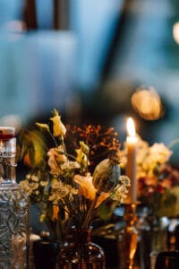 Eine sanft beleuchtete Szene mit einer brennenden Kerze, umgeben von Glasflaschen und Vasen, perfekt für eine Hochzeit. Die Vasen enthalten zarte, helle Blumen und Blätter und schaffen eine warme und gemütliche Atmosphäre. Der unscharfe Hintergrund suggeriert eine entspannte, intime Umgebung, ideal für Hochzeiten.