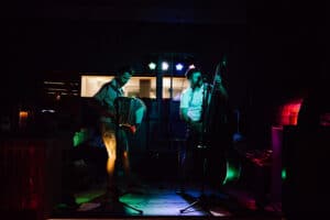 Unter farbenfrohen Bühnenlichtern treten zwei Musiker im Rahmen eines einzigartigen Hochzeitsplanungs-Events auf. Einer spielt Akkordeon, der andere Kontrabass. Ihre konzentrierten Ausdrücke werden durch den schwach beleuchteten Veranstaltungsort hervorgehoben und schaffen eine perfekte Atmosphäre für eine unvergessliche Hochzeit.